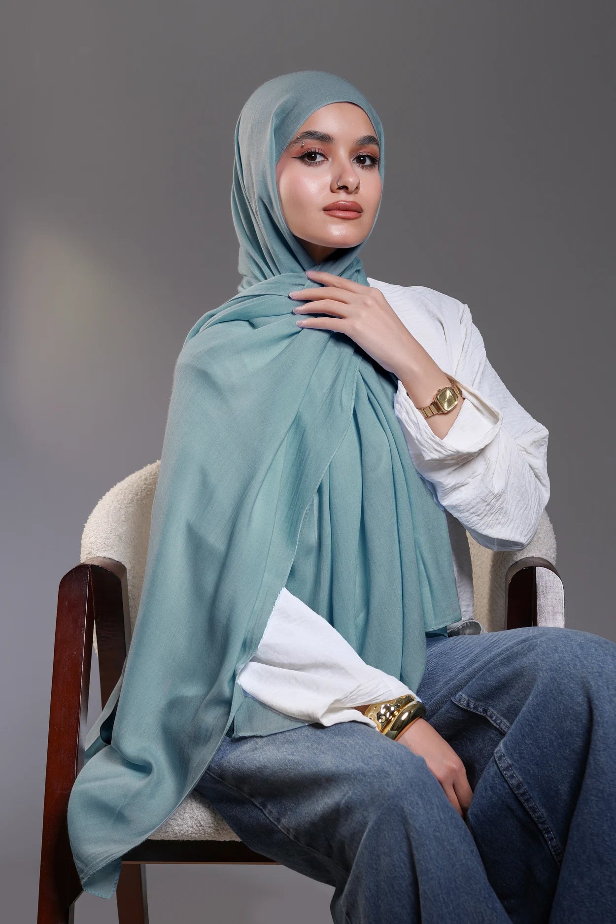Woman wearing a light blue hijab, white shirt, and blue jeans sitting on a chair against a gray background.
