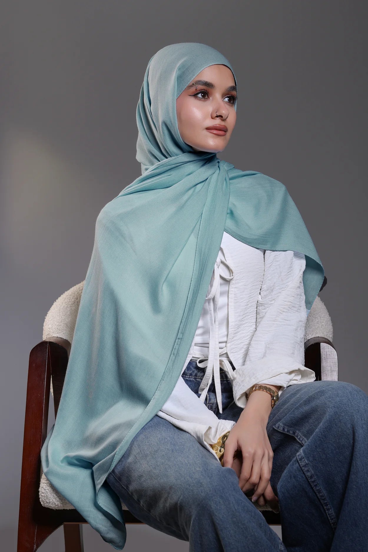 Woman wearing a light blue hijab, white shirt, and jeans sitting on a chair against a gray background
