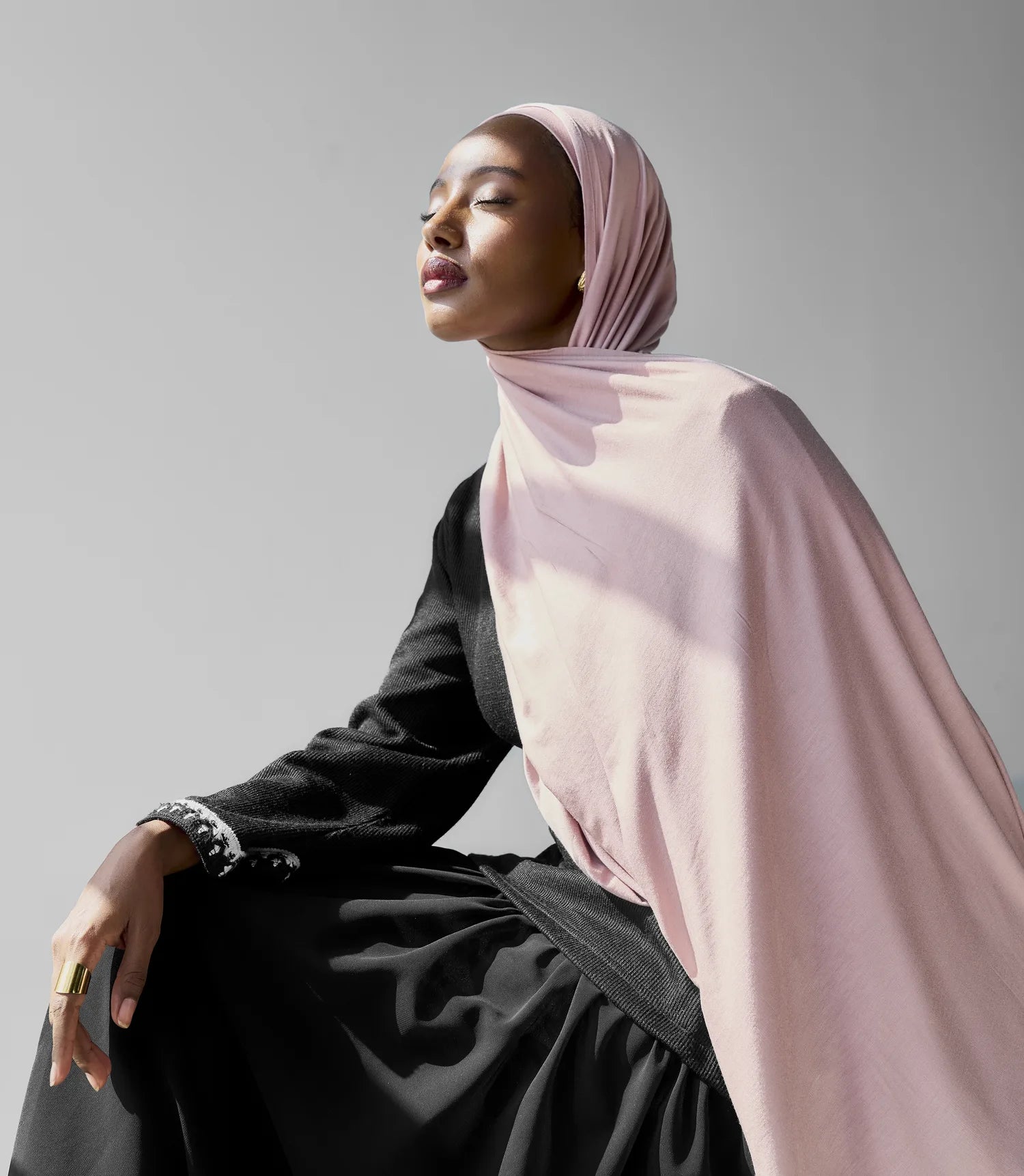 Woman wearing a light pink hijab and black outfit against a gray background