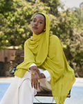 Woman wearing a bright yellow hijab sitting outdoors with greenery in the background