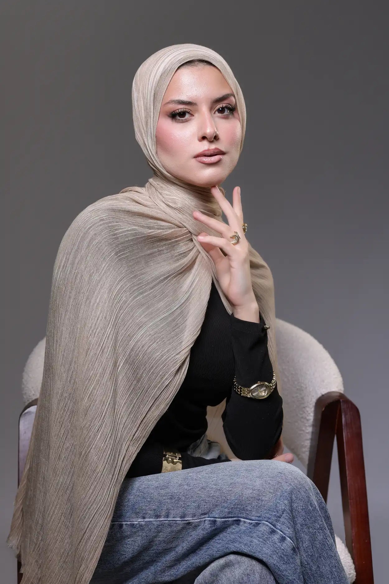 Woman wearing a beige hijab and black top sitting on a chair against a gray background