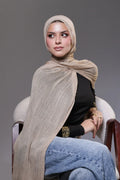 Woman wearing a beige hijab, black top, and blue jeans sitting on a chair against a gray background