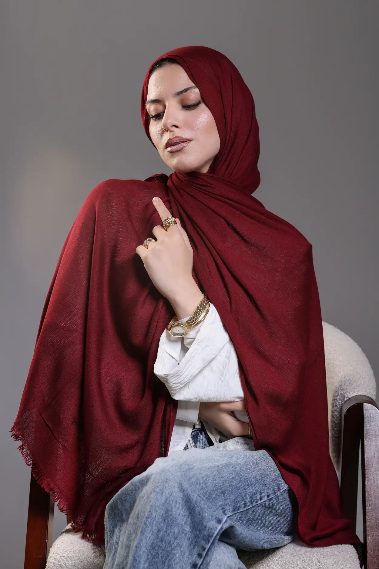 Woman wearing a red hijab and blue jeans sitting on a chair against a gray background