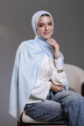 Woman wearing a light blue hijab and white top sitting on a chair against a gray background