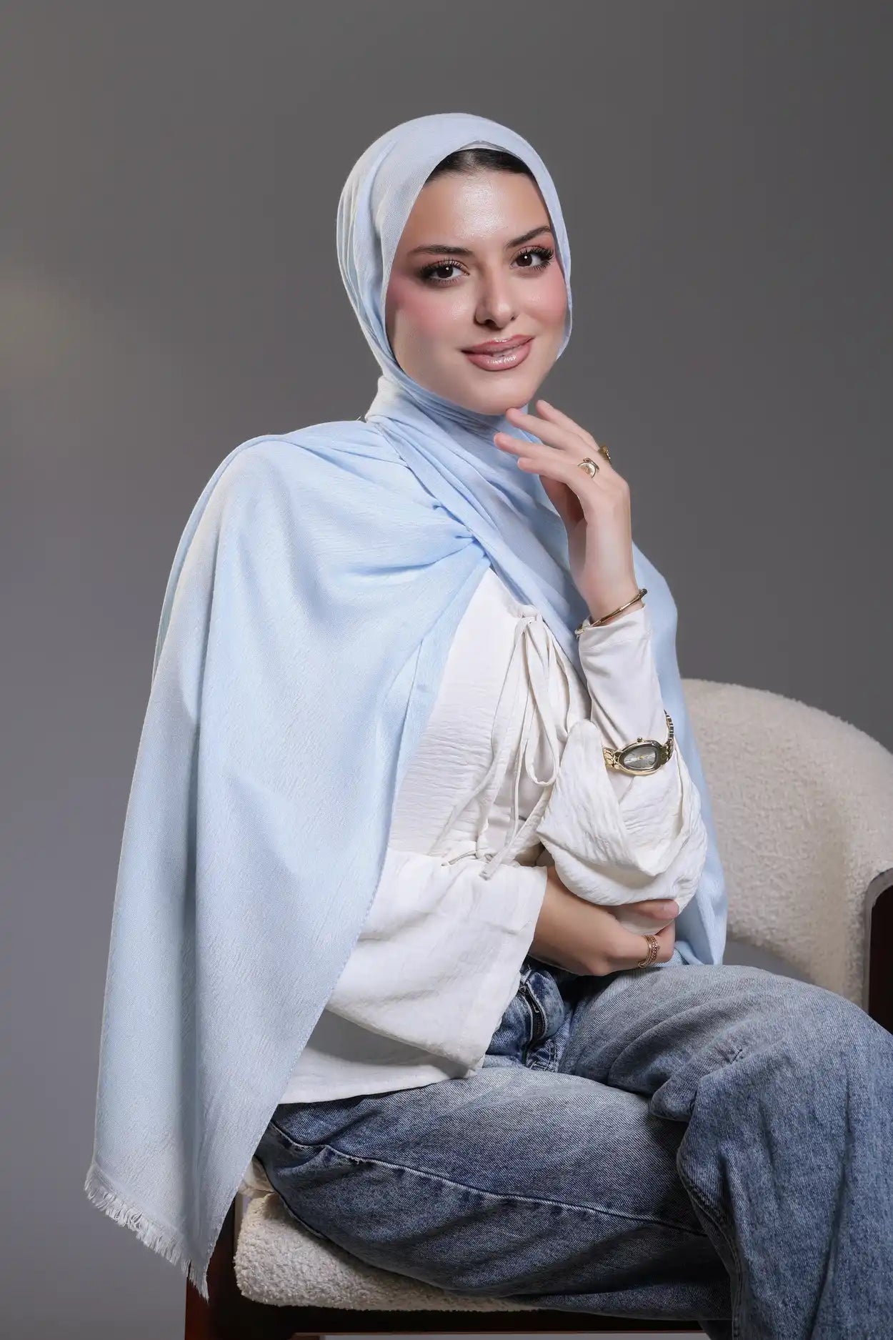 Woman wearing a light blue hijab and white top sitting on a chair against a gray background