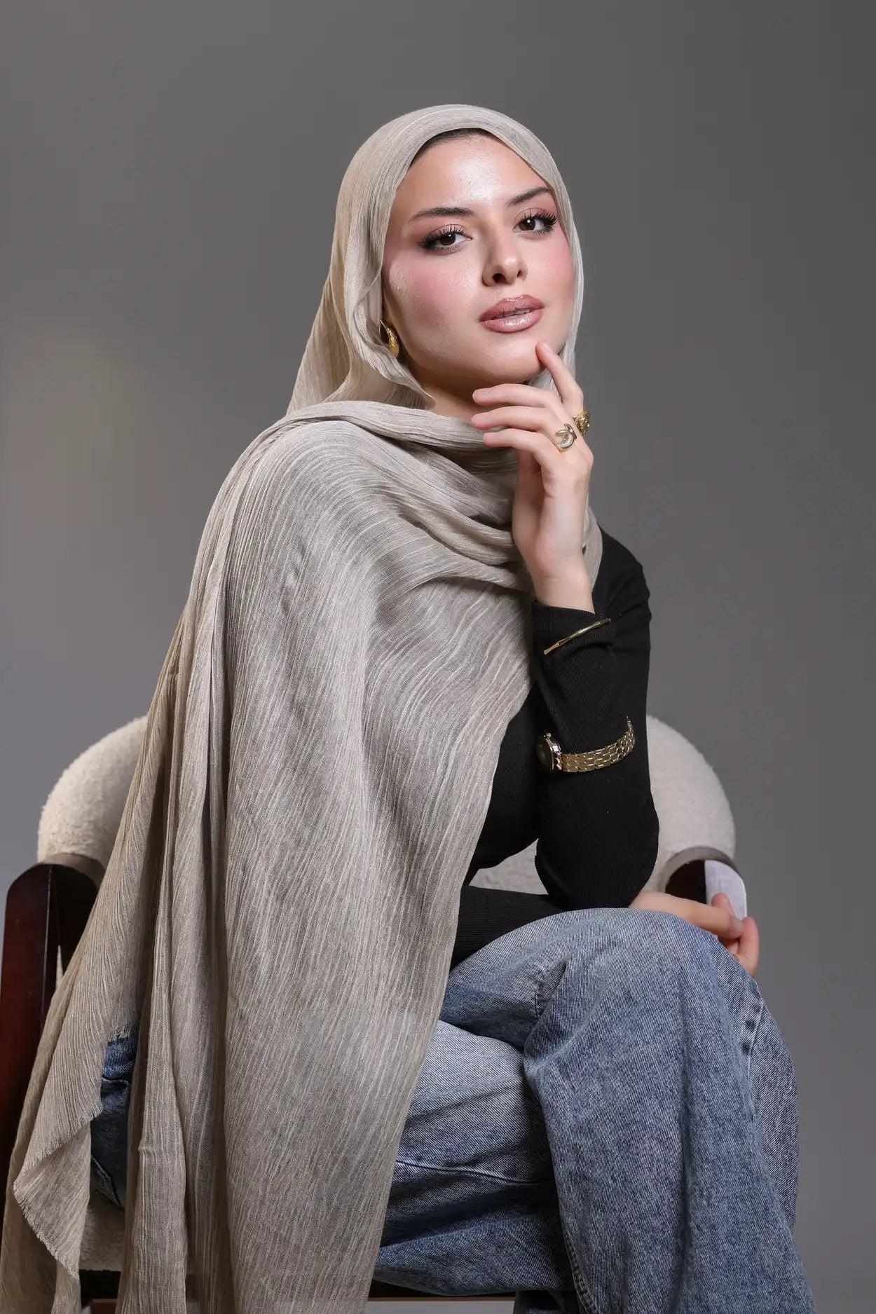 Woman wearing a beige hijab and black top sitting on a chair against a gray background