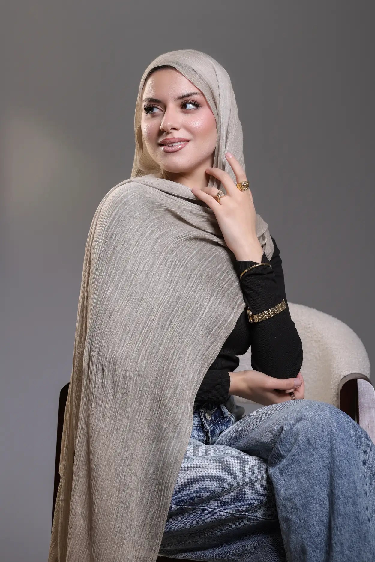 Woman wearing a beige hijab and black top, sitting on a chair against a gray background