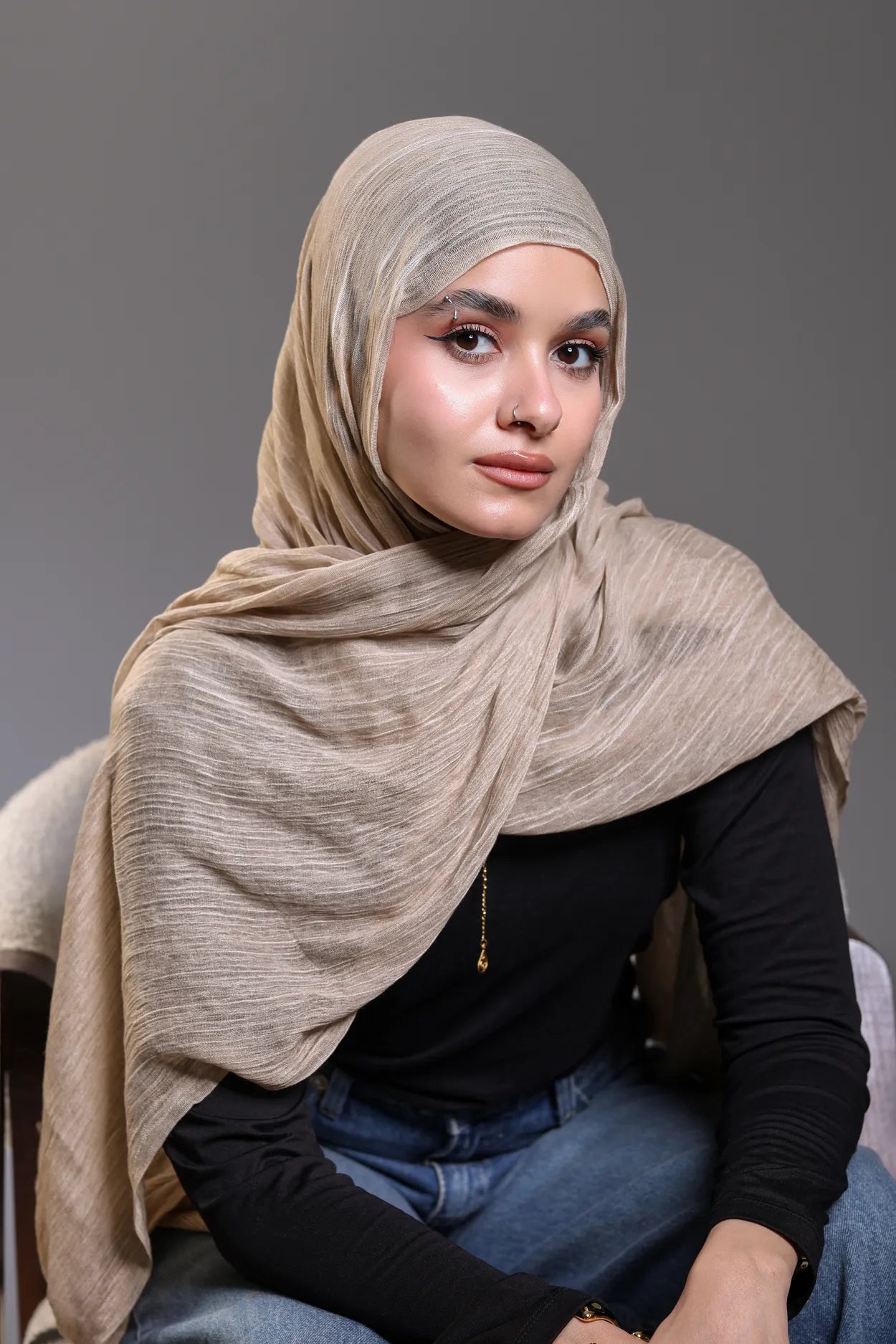 Woman wearing a beige hijab and black top against a gray background