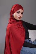 Woman wearing a red hijab, black top, and blue jeans against a gray background
