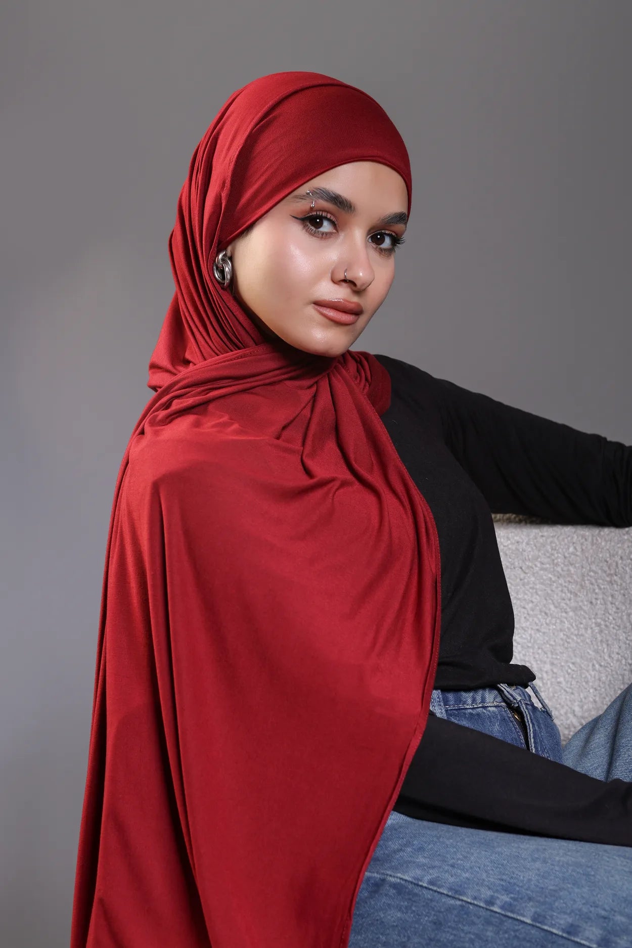 Woman wearing a red hijab, black top, and blue jeans against a gray background