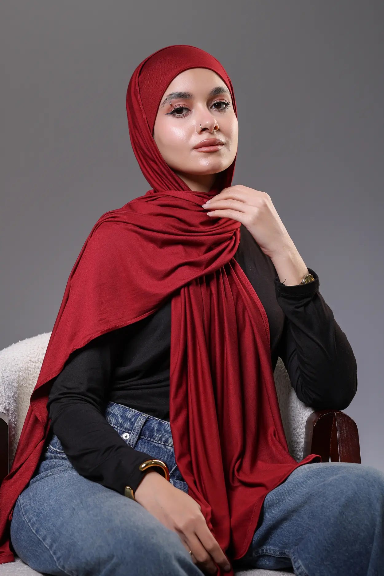 Woman wearing a red hijab, black top, and blue jeans sitting on a chair against a gray background