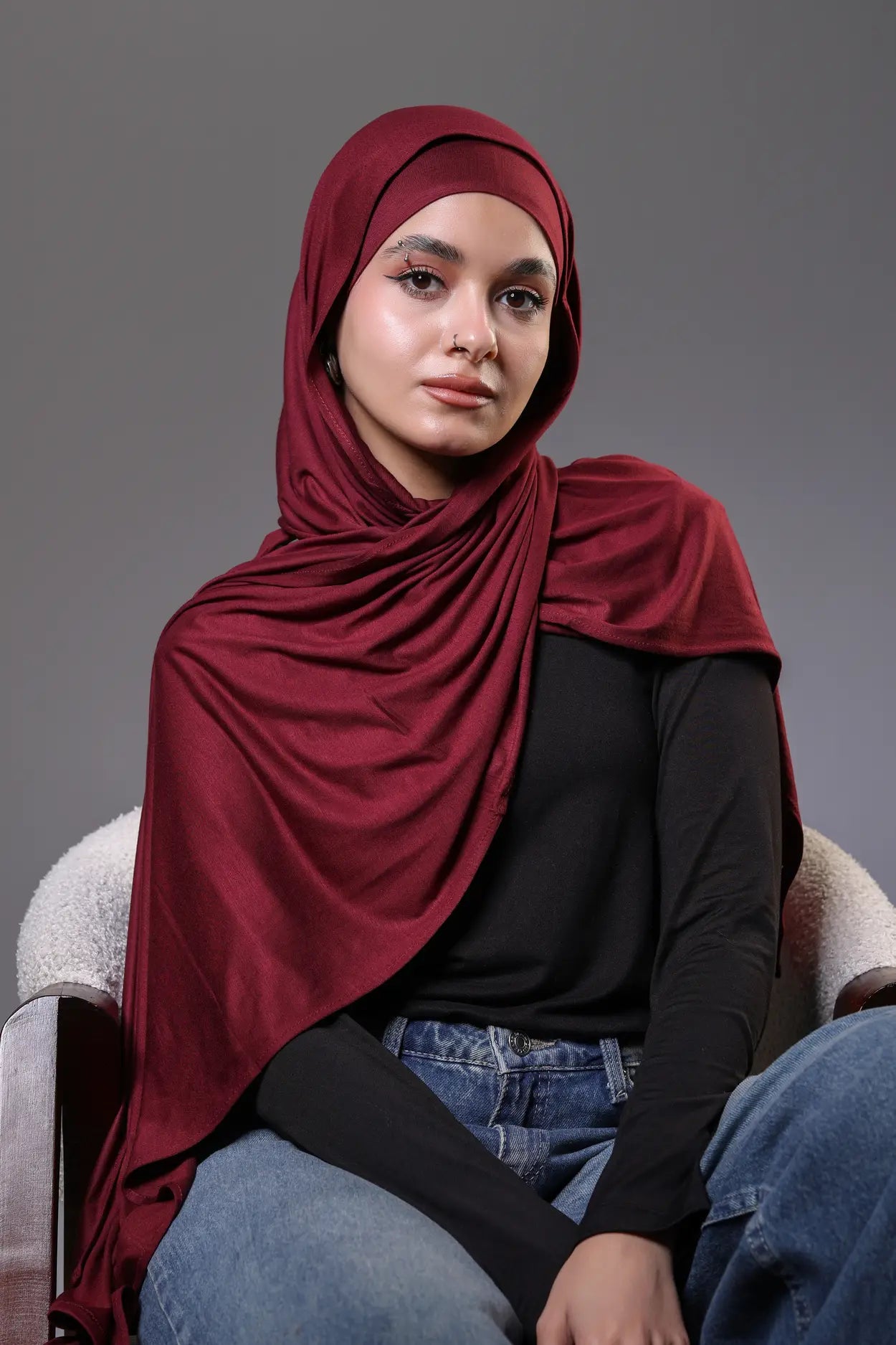 Woman wearing a red hijab and black top sitting on a chair against a gray background