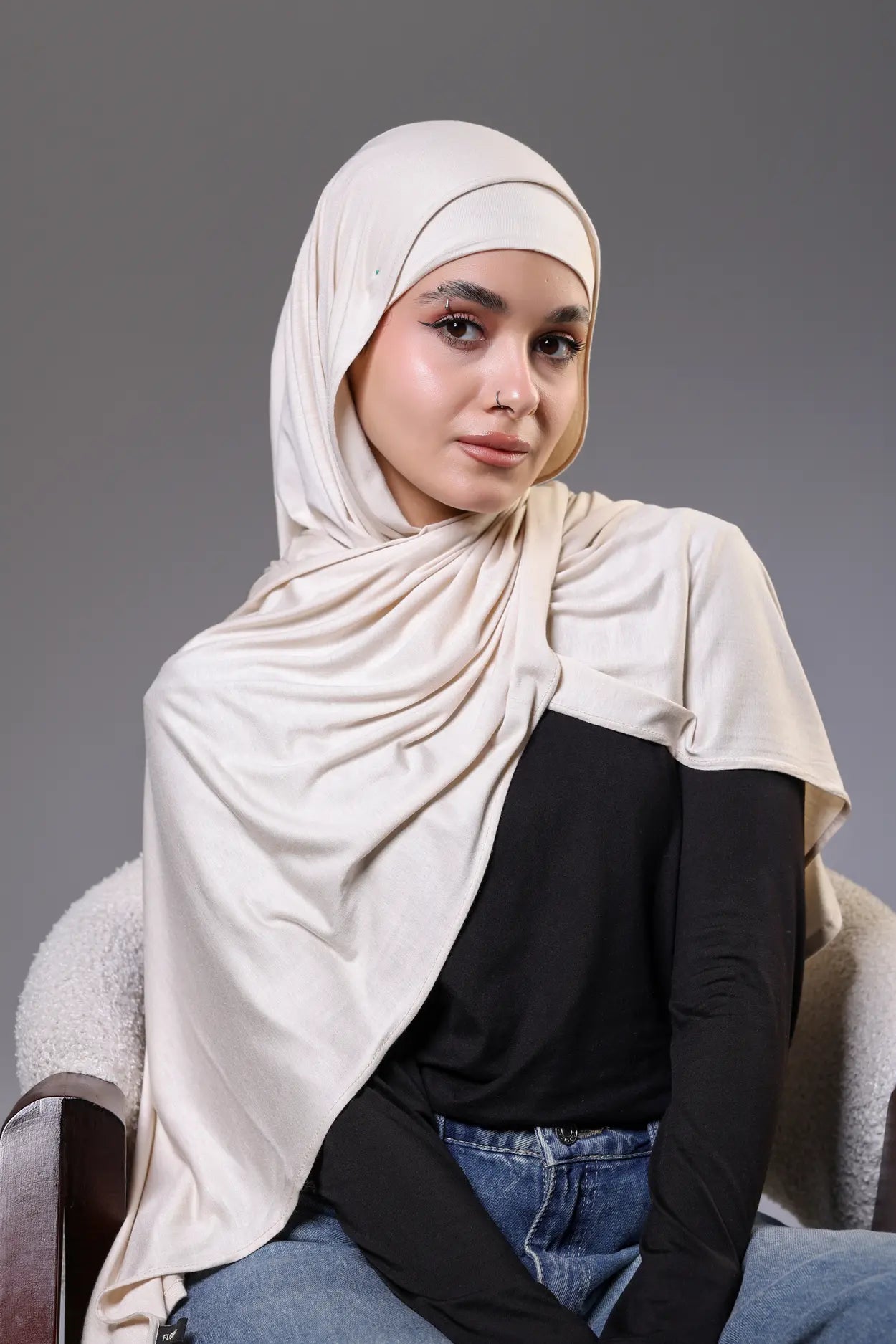 Woman wearing a beige hijab and black top against a gray background