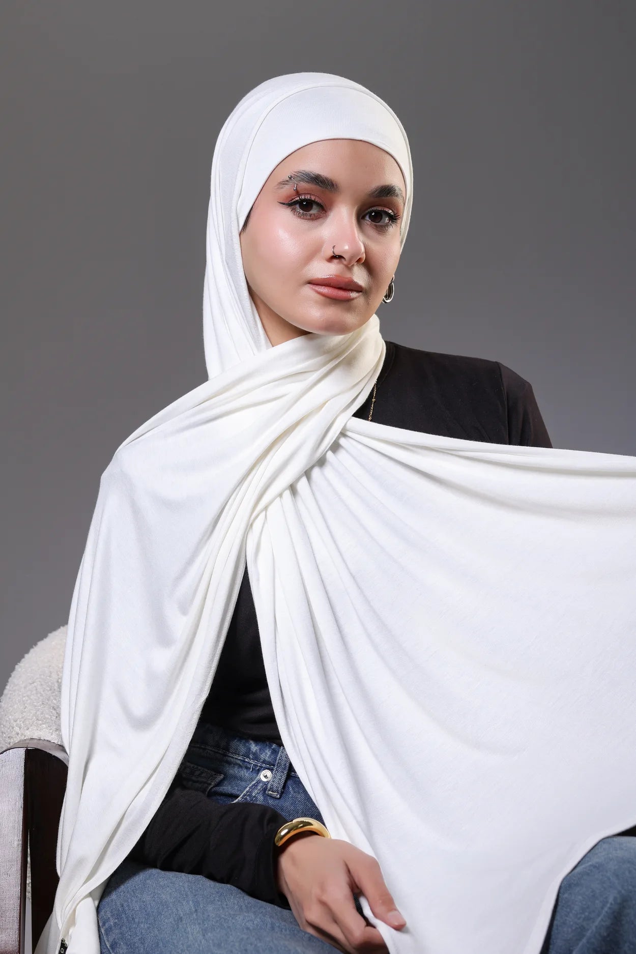 Woman wearing a white hijab and sitting on a chair against a gray background