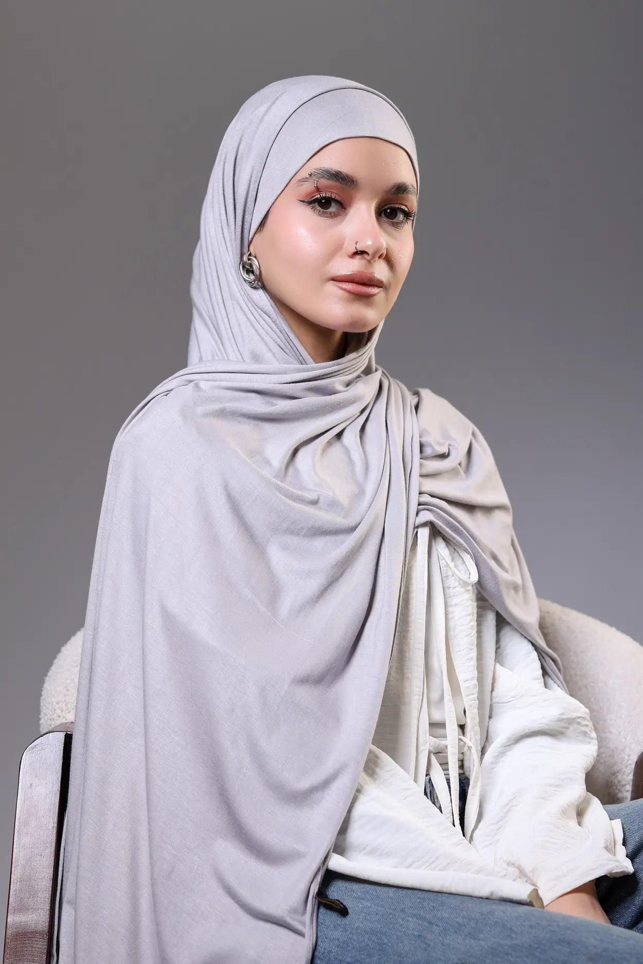 Woman wearing a light gray hijab and white top against a gray background